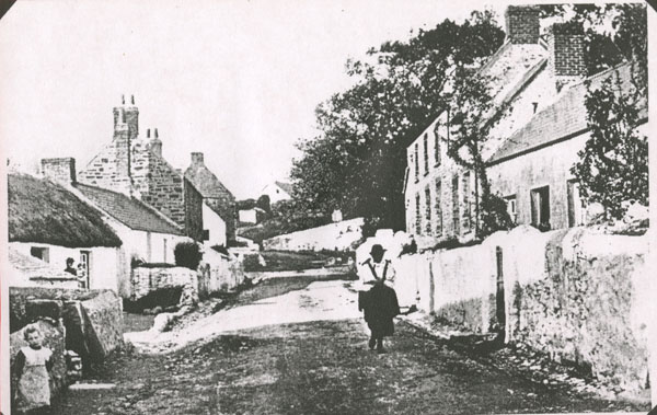 Copy of a photograph looking up Llangwm Pembrokeshire Main Street towards the shop and with the church on the left hand side. There is a lady walking towards the camera in traditional Llangwm costume and a little girl in the left of the photograph both unnamed